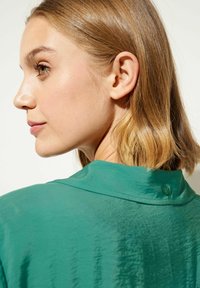 Green shirt with a soft, slightly shiny texture; features a collar and a small button detail at the back. Styled with medium-length hair.