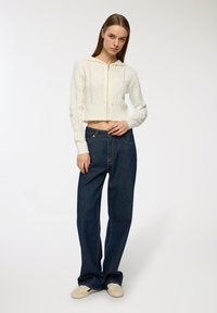 Przykrócony biały sweter w warkocz z kapturem, z diamentowym wzorem i ściągaczami na rękawach, w połączeniu z wysokim stanem ciemnoniebieskich dżinsów.