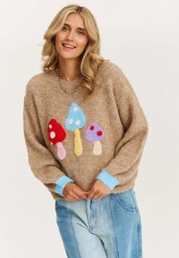 Beżowy sweter wykonany na drutach z kolorowymi motywami grzybów w odcieniach niebieskiego, czerwonego i różowego. Ma jasnoniebieskie mankiety i luźny krój.