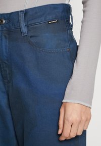 Modré džíny z denimu s jemným gradientem, které mají viditelné knoflíky, kapsu a značkovou etiketu. Materiál má hladkou texturu.