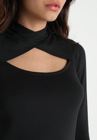 Top nero a maniche lunghe con colletto alto e design con scollatura sul petto. Tessuto liscio con leggero motivo a coste e dettaglio del logo.