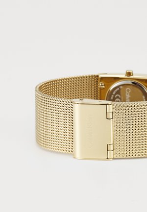 Bracelet de montre en maille dorée avec un boîtier carré et une boucle carrée. Dispose d'une texture lisse et d'un logo gravé sur la boucle.