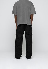 Person, der mit dem Rücken zur Kamera steht, trägt ein graues, übergroßes T-Shirt und schwarze Cargo-Hosen sowie weiße Sneakers vor einem schlichten Hintergrund.