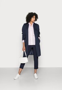 Trench coat blu navy con bottoni, camicia rosa, pantaloni cropped blu navy, borsa trapuntata bianca e sneaker bianche. Tessuto morbido, silhouette strutturata.