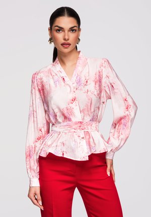 Femme aux cheveux noirs attachés en queue de cheval basse, portant un chemisier rose à fleurs avec une ceinture et un pantalon rouge vif, debout devant un fond clair.