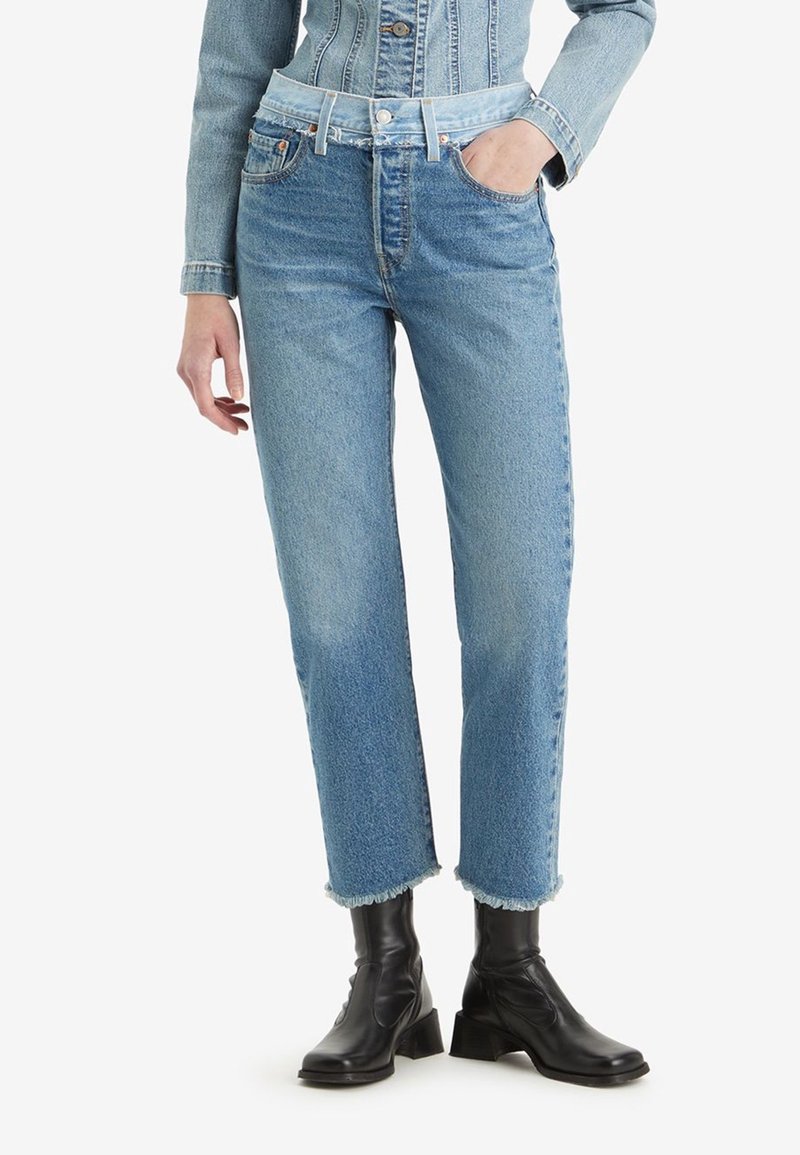 Femme portant un jean taille haute bleu clair, coupe droite avec ourlet brut et bottines noires, main dans la poche, debout sur fond blanc.
