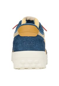 Scarpa blu in pelle scamosciata con tallone in cuoio marrone chiaro, linguetta beige e dettagli rossi. Presenta una suola bianca testurizzata e il logo "Hey Dude" sul retro.
