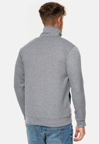 Grauer Reißverschluss-Sweatshirt mit hohem Kragen, glatter Textur, kontrastierenden Seitenpaneelen und gerippten Bündchen, getragen über blauen Jeans.