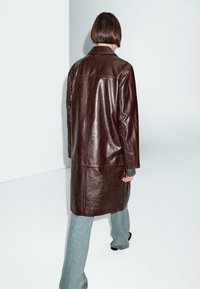 Cappotto lungo in pelle marrone con taglio dritto, dotato di colletto e dettagli di cucitura minimi, indossato sopra jeans grigi a vita alta.