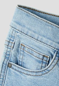 Kapsa na světlých modrých džínách s viditelným oranžovým šitím a malým kovovým nýtem v levém dolním rohu kapsy.