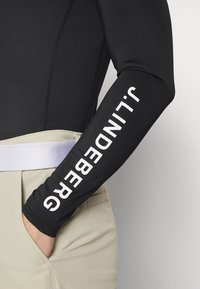 Crna majica dugih rukava s pripijenim dizajnom, s bijelim vertikalnim tekstom "J.LINDEBERG" na lijevom rukavu. Nosite je s bež hlačama visokog struka.