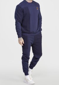 Conjunto de sudadera en azul marino con sudadera de cuello redondo texturizada y joggers a juego. Presenta un parche de logo y puños acanalados. El material parece suave.