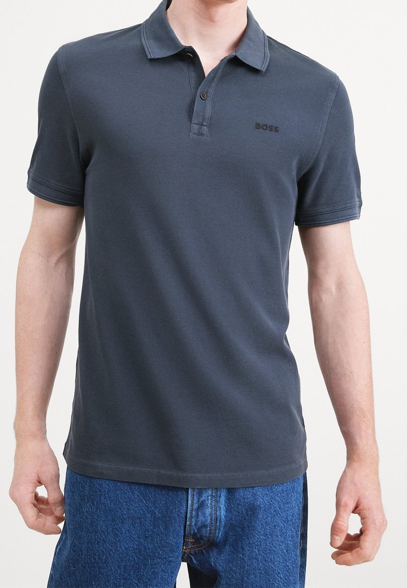 Homme portant un polo gris foncé à manches courtes avec un col et le logo "BOSS" sur la poitrine, associé à un jean bleu en denim.