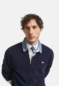 Tamsiai mėlyna polo marškiniai su ilgomis rankovėmis, šviesiai mėlynas tiesioginių apykaklė, mažas logotipas krūtinėje ir patikimų raštų kaklaraištis po juo.