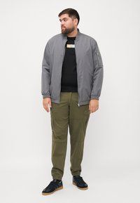 Grijze bomberjack, ritssluiting aan de voorkant, zwarte T-shirt eronder, olijfgroene cargobroek, zwarte sneakers met een gumrubber zool. Casual outfit ontwerp.