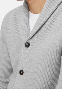 Grau gestrickte Strickjacke mit gerippter Textur, Schalkragen und zwei schwarzen Knöpfen auf der Vorderseite. Getragen über einem weißen Hemd.