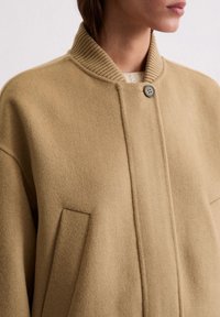 Beige Wolljacke mit geripptem Kragen, einem einzigen Knopfverschluss und zwei seitlichen Taschen. Weicher Stoff und minimalistisches Design.