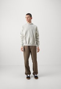 Hanorac gri cu buzunar frontal, asortat cu pantaloni maro și adidași negri cu șireturi albe. Textură netedă și croială lejeră.