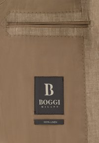 Tissu beige avec une doublure cousue, une étiquette noire indiquant « B Boggi Milano » et une petite étiquette mentionnant « 100 % lin ».