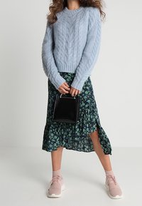 Frau in hellblauem Kabelstrickpullover, floralem Midikleid mit seitlichem Schlitz, rosa Sneakers, eine kleine schwarze Handtasche haltend vor weißem Hintergrund.