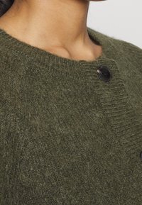 Olivgrön kofta gjord av mjukt, strukturerat tyg, med rund hals och svarta knappar. Stickningen har ett subtilt randigt mönster.