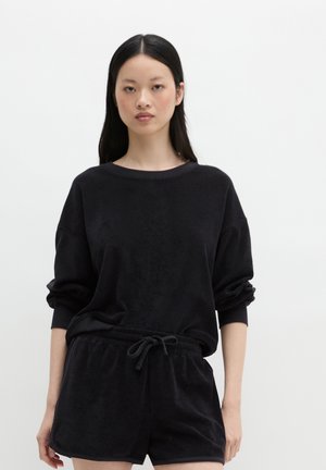 Młoda kobieta z długimi czarnymi włosami, ubrana w czarny top z długim rękawem i pasujące czarne szorty na jednolitym białym tle.