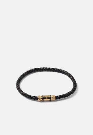 Bracelet en cuir tressé noir avec un fermoir magnétique cylindrique en or comportant le nom de la marque gravé sur fond blanc.