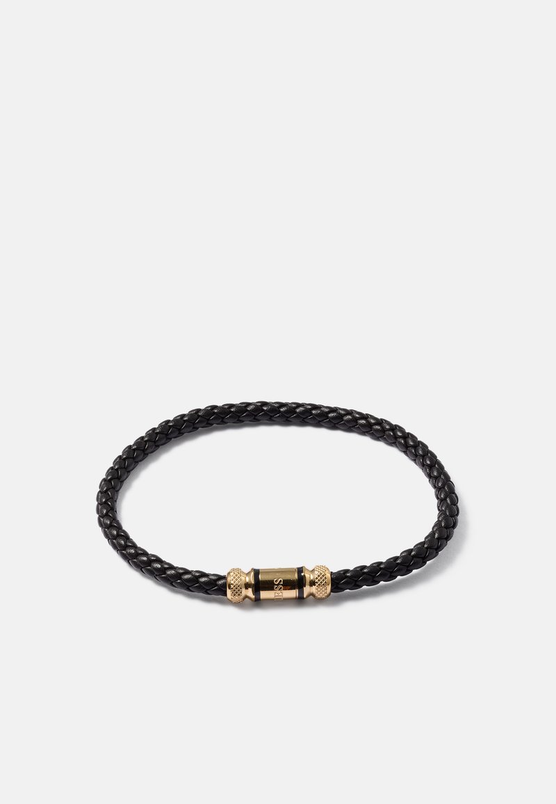 Bracelet en cuir tressé noir avec un fermoir magnétique cylindrique en or comportant le nom de la marque gravé sur fond blanc.