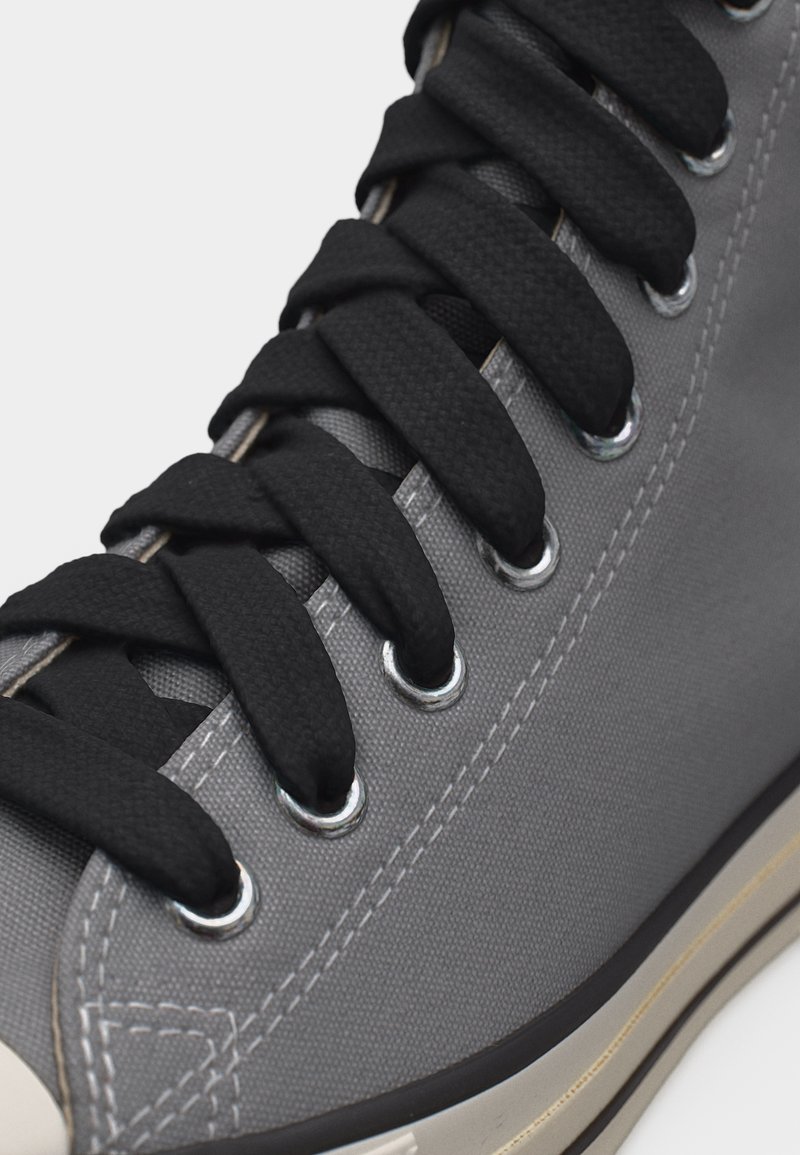 Primo piano di una sneaker grigia con lacci neri infilati attraverso occhielli metallici e cuciture bianche visibili sul tessuto superiore.