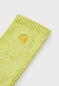 Grüne, gerippte Socke mit einem gelben, gestickten Logo. Das Material wirkt weich mit einer strukturierten Oberfläche und einem eng sitzenden Bündchen.