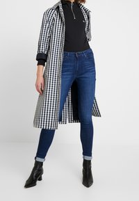Persona que lleva unos pantalones skinny de mezclilla azul oscuro, botas negras hasta el tobillo, una chaqueta negra con cremallera y un abrigo largo a cuadros en blanco y negro abierto.