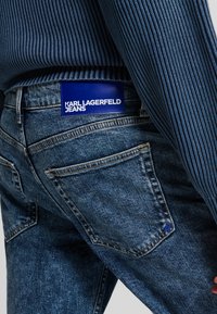 Jeans en denim dans une teinte bleue moyenne avec une texture usée, arborant une étiquette bleue sur la taille portant la mention "KARL LAGERFELD JEANS."