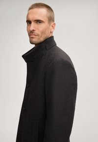 Manteau en laine mélangée noir avec un col haut, des boutons croisés et des poches latérales. Texture en chevrons, forme structurée et coupe ajustée.