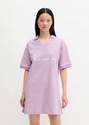 Jonge vrouw met lang zwart haar die een losse lila polojurk draagt met de tekst "It's on us." op de voorkant gedrukt.