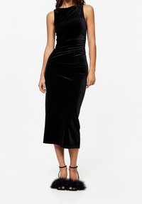 Robe midi en velours noir avec une silhouette ajustée, des détails froncés et un col rond. Associée à des sandales noires à brides avec des accents en fausse fourrure.