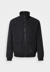 Sort lynlåsjakke med høj krave, to forlommer med klap, ribbede manchetter og kant samt lille logo-patch nederst til højre.