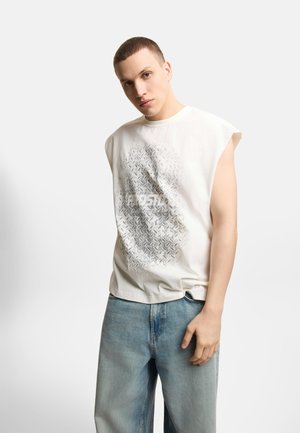 Młody mężczyzna z krótkimi włosami, ubrany w białą koszulkę bez rękawów z nadrukiem i jasnoniebieskie dżinsy, stoi na jednolitym białym tle.