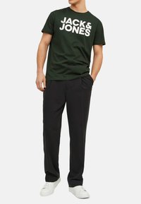 Dark green cotton t-shirt with large white "JACK & JONES" lettering, paired with black trousers and white sneakers. Minimal design, casual fit.