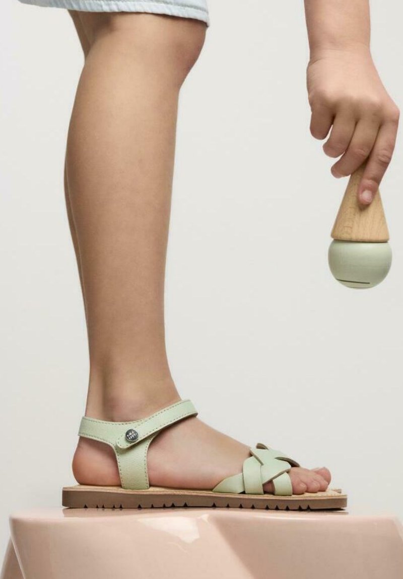 Sandal in pelle verde chiaro con un design a fascia incrociata e dettaglio in metallo circolare. Suola in gomma marrone con motivo di battistrada testurizzato.