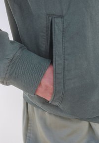 Grüne Stoffjacke mit gerippten Bündchen und einer Seitentasche. Die Textur ist glatt, mit genähten Details und einem lässigen Design.