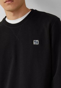 Uomo che indossa un maglione nero a girocollo con un piccolo patch con il logo "s.O" sul petto, sovrapposto a una maglietta bianca.