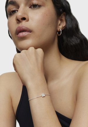 Pulsera de cadena de plata con un acento de piedra clara y con forma de ondas. La modelo lleva una blusa sin mangas de color oscuro, mostrando la pulsera en su muñeca.