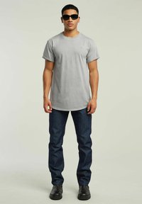 Szara koszulka z okrągłym dekoltem i podwiniętymi rękawami, zestawiona z ciemnoniebieskimi dżinsami i czarnymi butami. Model nosi czarne okulary przeciwsłoneczne.