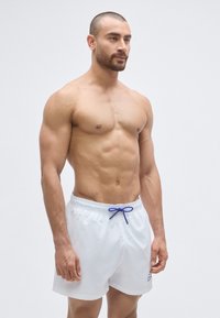 Fehér fürdőnadrág könnyű anyagból, rugalmas derékrésszel és kék zsinórral. Nincs rajta minta vagy egyéb díszítés.