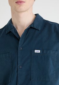 Navy blue short-sleeve shirt with a button front, two pockets, and a "Lee" brand label on the left pocket. Smooth, woven fabric texture.