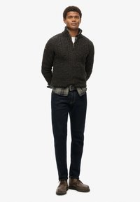 Dunkler gestrickter Pullover mit hohem Kragen, über einem karierten Hemd getragen, kombiniert mit dunklen Jeans und braunen Schnürstiefeln.