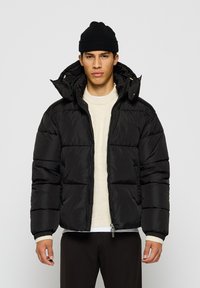 Ung mann iført svart beanie, svart puffjakke over kremfarget genser, står mot en ensfarget lys bakgrunn med ansiktet vendt fremover.