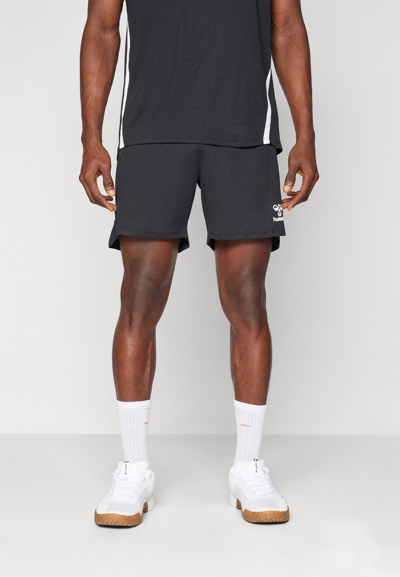 Hombre vestido con pantalones cortos deportivos negros, camiseta deportiva en blanco y negro, calcetines deportivos blancos y zapatillas blancas, de pie sobre un fondo liso.
