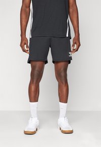 Hombre vestido con pantalones cortos deportivos negros, camiseta deportiva en blanco y negro, calcetines deportivos blancos y zapatillas blancas, de pie sobre un fondo liso.