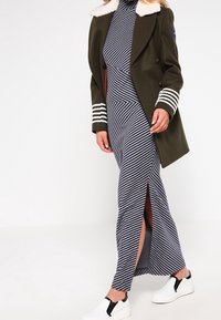Abrigo verde olivo de doble botonadura con un cuello de piel blanca, combinado con un vestido maxi a rayas azul marino y blanco con aberturas laterales, y acompañado de zapatillas blancas.